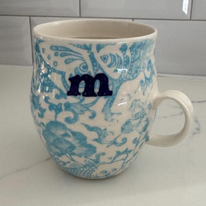 Puede incluir: Taza de cer&aacute;mica con base crema y estampado floral azul. La taza tiene la letra "m" en azul oscuro. El asa es de dise&ntilde;o simple y curvado. Adecuada para bebidas fr&iacute;as o calientes. Altura aproximada de 10 cm.