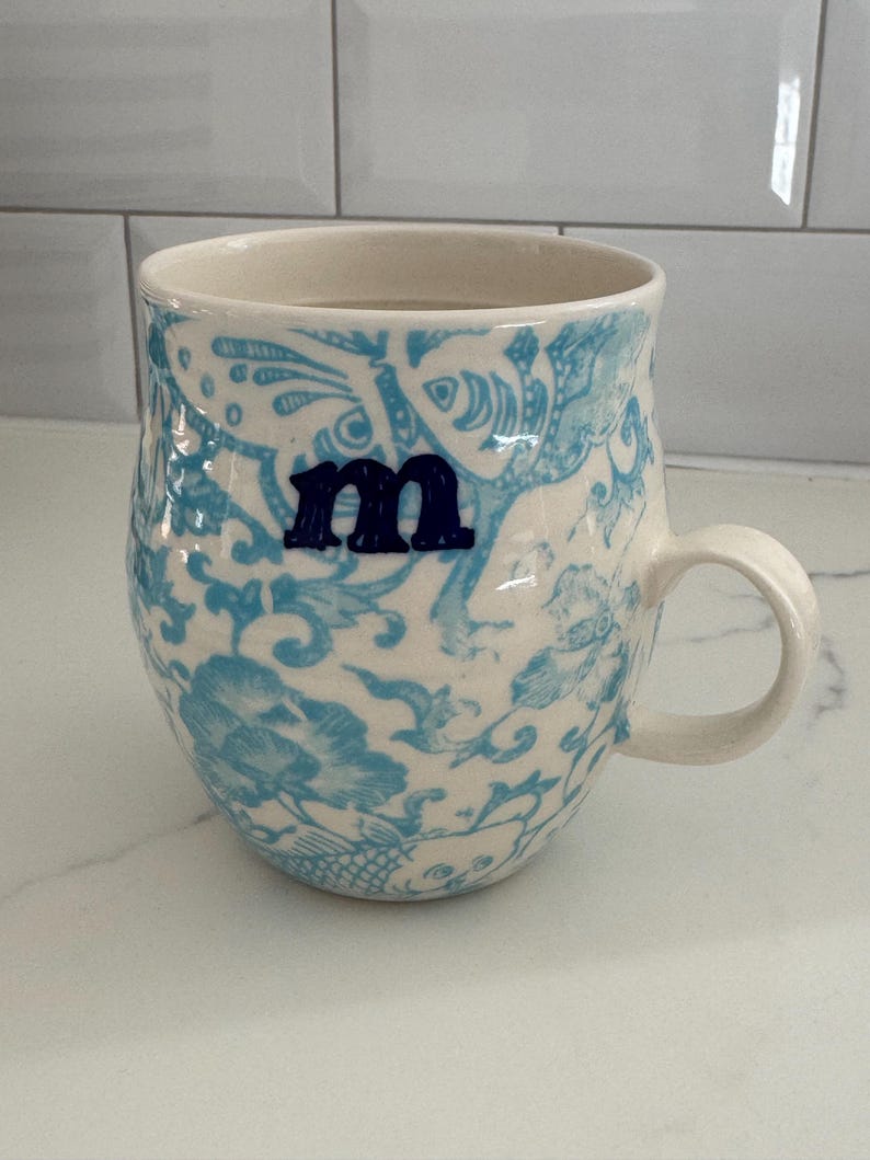 Puede incluir: Taza de cer&aacute;mica con base crema y estampado floral azul. La taza tiene la letra "m" en azul oscuro. El asa es de dise&ntilde;o simple y curvado. Adecuada para bebidas fr&iacute;as o calientes. Altura aproximada de 10 cm.