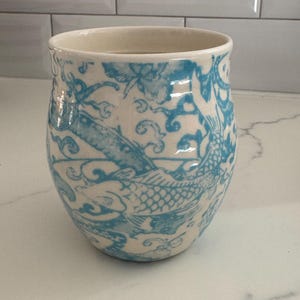 Puede incluir: Un vaso de cer&aacute;mica con interior y exterior color crema. El exterior presenta un dise&ntilde;o de pez y flores azules. El vaso tiene forma redondeada y una abertura ancha. El dise&ntilde;o recuerda al arte asi&aacute;tico tradicional.