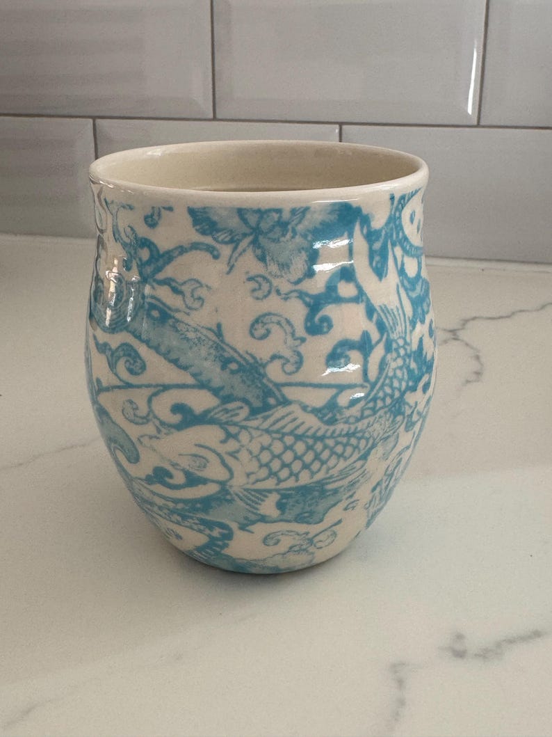 Puede incluir: Un vaso de cer&aacute;mica con interior y exterior color crema. El exterior presenta un dise&ntilde;o de pez y flores azules. El vaso tiene forma redondeada y una abertura ancha. El dise&ntilde;o recuerda al arte asi&aacute;tico tradicional.