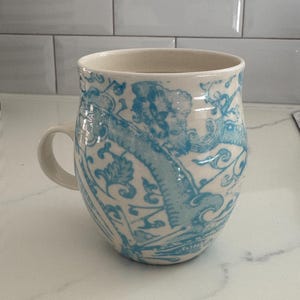 Puede incluir: Una taza de cer&aacute;mica con interior y asa color crema. El exterior presenta un dise&ntilde;o azul claro adornado con motivos florales y de volutas. La taza est&aacute; sobre una encimera blanca, mostrando su detallada artesan&iacute;a.