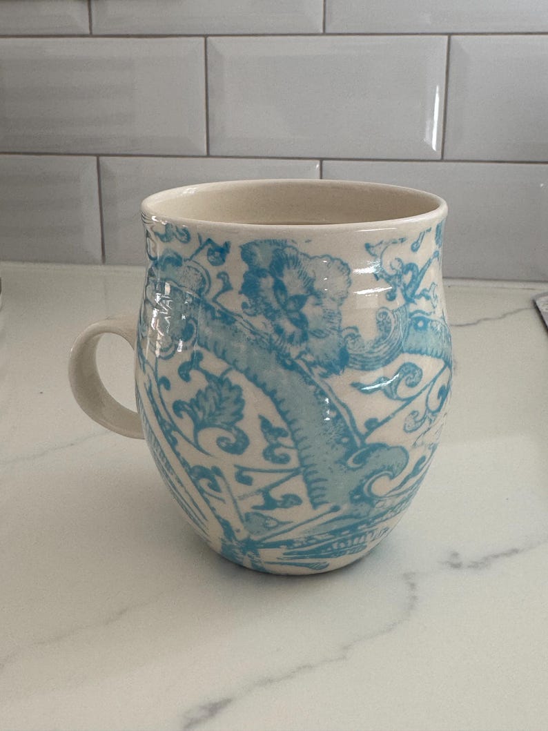 Puede incluir: Una taza de cer&aacute;mica con interior y asa color crema. El exterior presenta un dise&ntilde;o azul claro adornado con motivos florales y de volutas. La taza est&aacute; sobre una encimera blanca, mostrando su detallada artesan&iacute;a.
