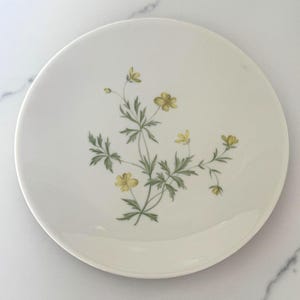 May include: A white ceramic plate featuring a delicate floral design. The design includes yellow flowers and green leaves arranged in a naturalistic pattern. The plate has a simple, elegant design, suitable for serving food.