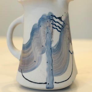 May include: A white ceramic pitcher with a handle and spout. The pitcher is decorated with blue and gray abstract designs, including wavy lines and a vertical element. A black line curves along the bottom.