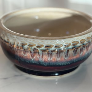 May include: A ceramic bowl with a glossy finish. The bowl features a color gradient, transitioning from a dark burgundy base to a light beige rim. The rim has a textured pattern with brown and gold accents. The bowl is round and sits on a small base.