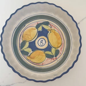 May include: A round, ceramic plate with a scalloped edge. The plate features a hand-painted design of yellow lemons, green leaves, and a blue and white border. The center has a blue background with a white swirl.