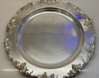 May include: A silver-colored, round serving tray with a decorative grape vine design around the rim. The tray has a reflective surface and a slightly raised edge. The grape vine design is detailed and adds an elegant touch to the tray.