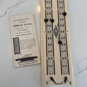 May include: A vintage pegless cribbage board with a cream-colored surface and black sliding markers. The board features two scoring tracks with numbered increments and a diamond-shaped logo. A small instruction booklet is included, with text that reads "Cribbage Board".