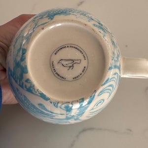 Puede incluir: Una taza de cer&aacute;mica blanca con un dise&ntilde;o floral azul. La base de la taza tiene el texto "MICROWAVE & DISHWASHER SAFE", un logotipo de p&aacute;jaro y "MADE IN CHINA". La taza tiene un asa.