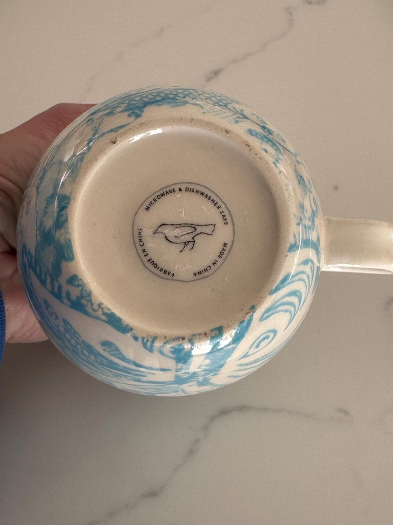 Puede incluir: Una taza de cer&aacute;mica blanca con un dise&ntilde;o floral azul. La base de la taza tiene el texto "MICROWAVE & DISHWASHER SAFE", un logotipo de p&aacute;jaro y "MADE IN CHINA". La taza tiene un asa.