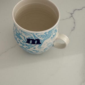 Puede incluir: Una taza de cer&aacute;mica con interior y asa de color crema. El exterior presenta un patr&oacute;n azul y blanco con la letra "m" en azul marino. La taza est&aacute; sobre una superficie de m&aacute;rmol blanco.