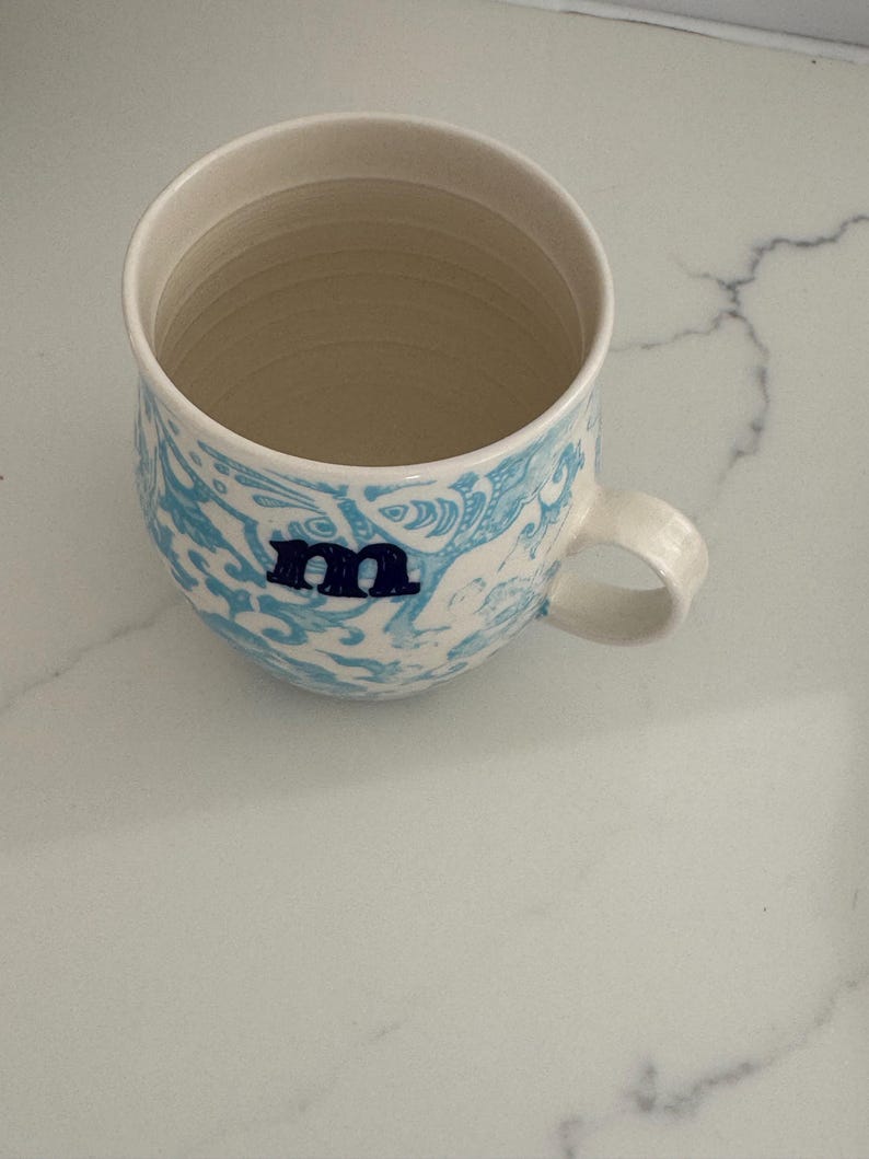Puede incluir: Una taza de cer&aacute;mica con interior y asa de color crema. El exterior presenta un patr&oacute;n azul y blanco con la letra "m" en azul marino. La taza est&aacute; sobre una superficie de m&aacute;rmol blanco.