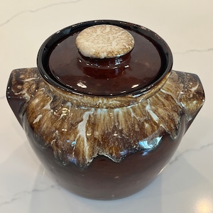 May include: A brown and white glazed ceramic cookie jar with a lid. The jar has a rounded body and two handles. The lid has a white knob.