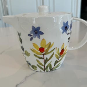 May include: White ceramic teapot with a floral design featuring yellow, blue, and green flowers. The teapot has a lid and a handle.