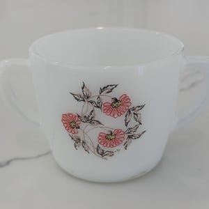 May include: White glass teacup with two handles, decorated with a floral design. The flowers are pink with yellow centers, surrounded by green and brown leaves. The teacup is a vintage style.