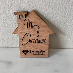 Puede incluir: Adorno de madera con forma de casa con el texto "Merry Christmas" y un logotipo para "Systematic Home Staging" que presenta un diamante.