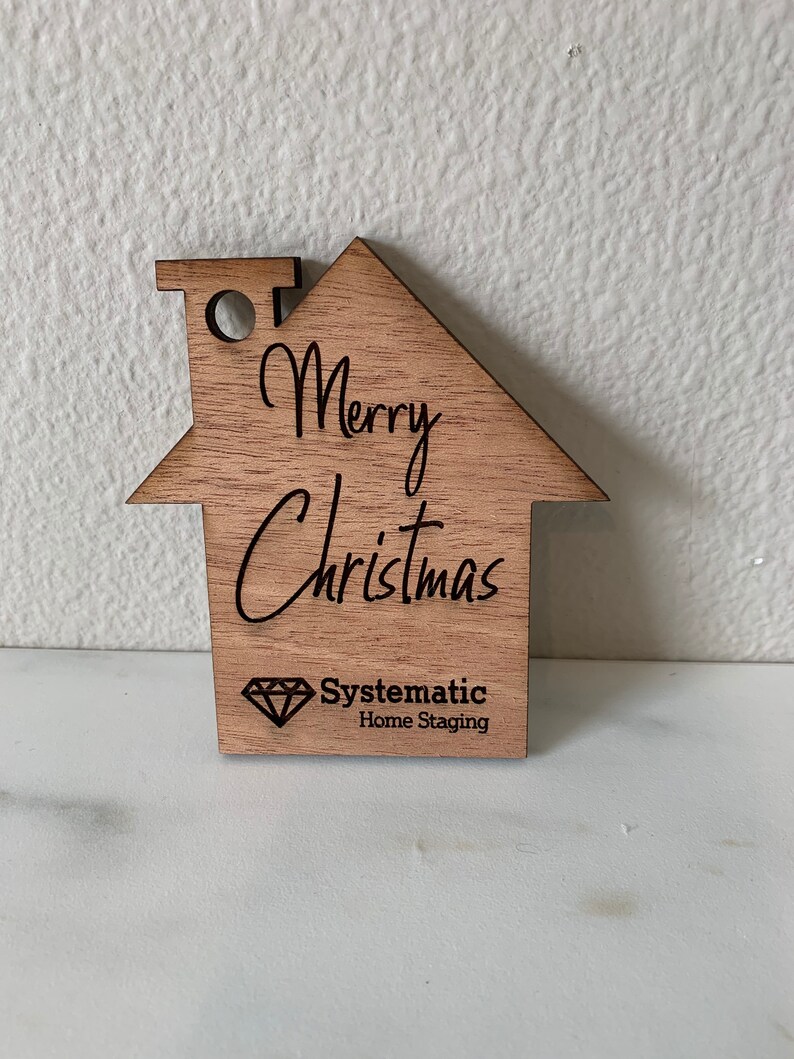 Puede incluir: Adorno de madera con forma de casa con el texto "Merry Christmas" y un logotipo para "Systematic Home Staging" que presenta un diamante.