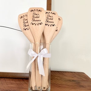 以下が含まれることがあります： 「Elise's Bridal Shower」と刻まれた木製のスプーンのセット。スプーンは透明なガラスの花瓶に白いリボンで飾られています。