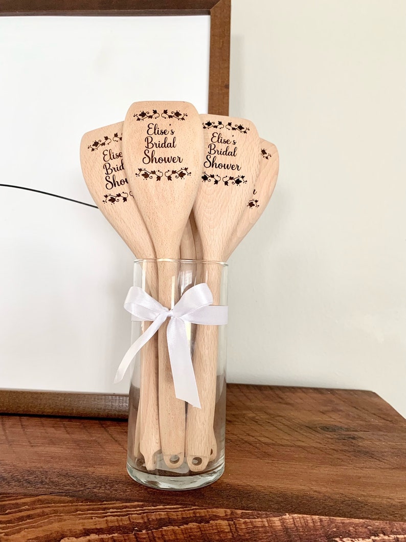以下が含まれることがあります： 「Elise's Bridal Shower」と刻まれた木製のスプーンのセット。スプーンは透明なガラスの花瓶に白いリボンで飾られています。