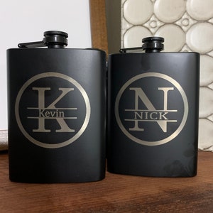 May include: Two black metal hip flasks with personalized engravings. The flask on the left has a circle with the letter K and the name Kevin. The flask on the right has a circle with the letter N and the name Nick.