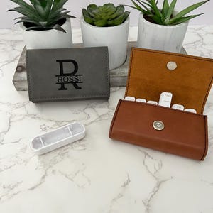 May include: A gray leather pill organizer with the initial "R" and the name "ROSSI" engraved on the front. The organizer is open to reveal seven compartments labeled with the days of the week. A brown leather pill organizer is also shown closed.