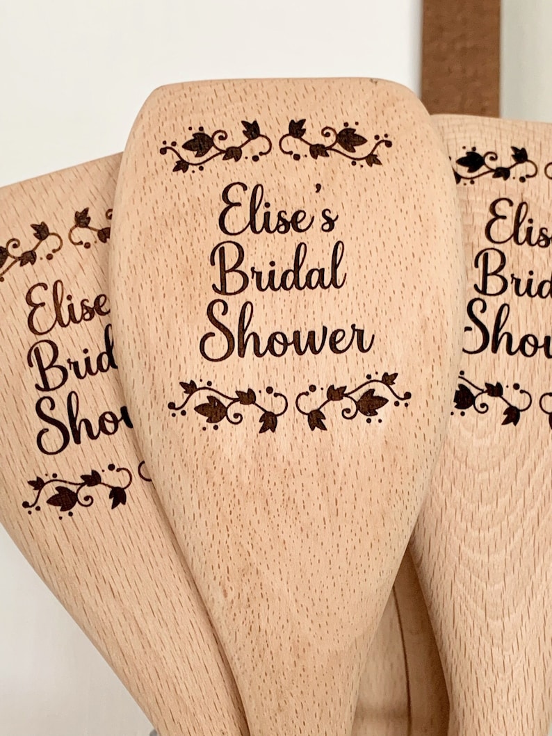 以下が含まれることがあります： 「Elise's Bridal Shower」と刻まれた木製のスプーン3本。スプーンはナチュラルな木製の仕上げで、シンプルな花の模様が施されています。