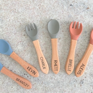 May include: A set of six baby utensils: three forks and three spoons. The fork and spoon heads are in blue, gray, and coral colors. The wooden handles are engraved with names: Brantley, Kevin, Charlie, Rhett, Brielle, and Alexis.