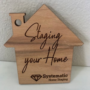 Puede incluir: Un letrero de madera con forma de casa con el texto "Staging your Home" y el logotipo "Systematic Home Staging" en forma de diamante.