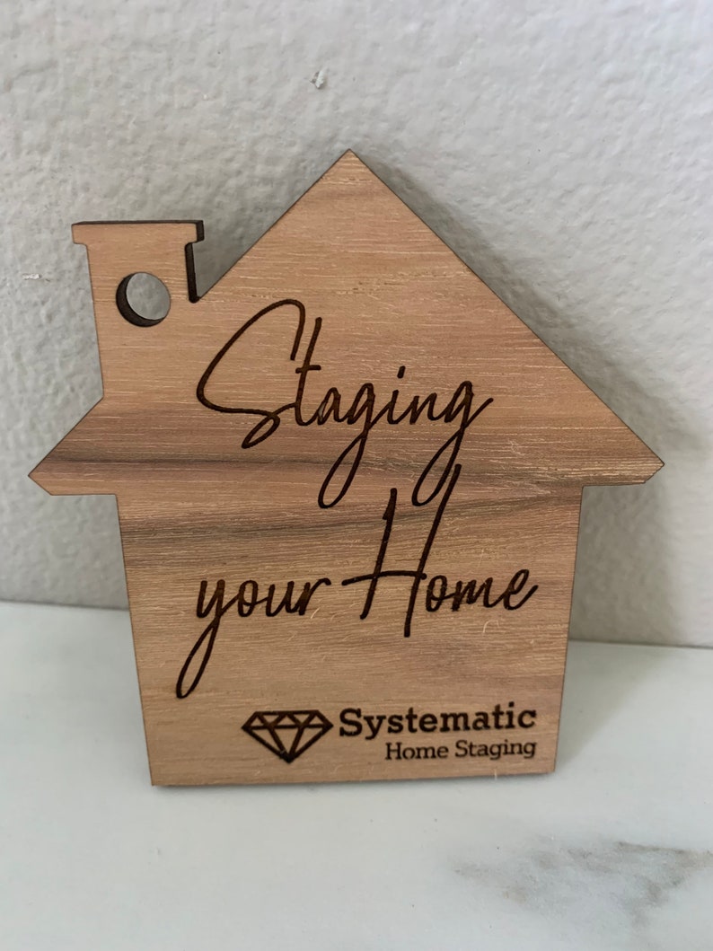 Puede incluir: Un letrero de madera con forma de casa con el texto "Staging your Home" y el logotipo "Systematic Home Staging" en forma de diamante.