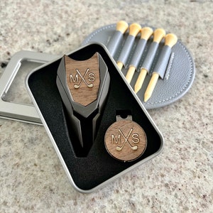 May include: A silver metal golf ball marker and a golf divot tool in a silver tin with a black interior. The golf ball marker and divot tool are both engraved with the initials "M.X.S." and a pair of crossed golf clubs.