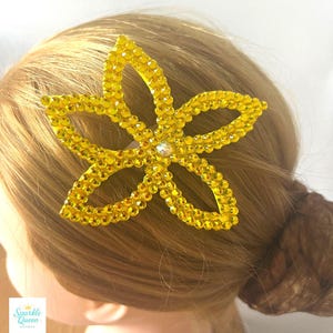 Op de afbeelding: Een gele, bloemvormige haarclip versierd met sprankelende strass-steentjes. De clip heeft een centrale, heldere strass-steen. Het haaraccessoire is bevestigd aan blond haar, gestyled in een knot. Het logo "Sparkle Queen Designs" is zichtbaar.
