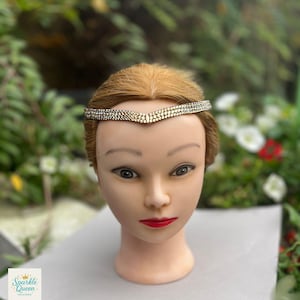 Op de afbeelding: Een zilveren strass-hoofdband met een centraal V-vormig ontwerp wordt getoond op een mannequinhoofd. De hoofdband schittert. De mannequin heeft blond haar, rode lippenstift en staat tegen een vage groene en witte bloemenachtergrond. Het logo "Sparkle Queen Designs" staat linksonder.