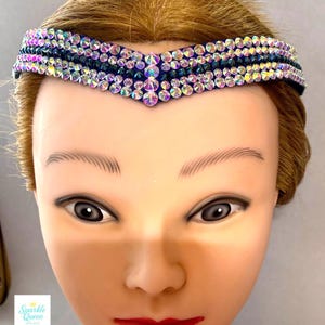 May include: A jewelled headband with a dark blue base and rows of iridescent rhinestones. The headband has a V-shaped design in the centre. The image also shows a mannequin head with the text "Sparkle Queen Designs".