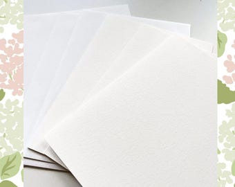May include: A stack of white watercolor paper sheets, fanned out to display the different textures and shades. The paper has a slightly textured surface, ideal for painting and drawing. The background features a floral pattern in shades of green and pink.