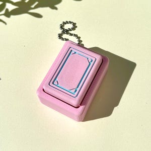 May include: A pink rectangular keychain with a light blue rectangular design on the top. The keychain is attached to a silver chain. The background is a light yellow color with green leaves and shadows.