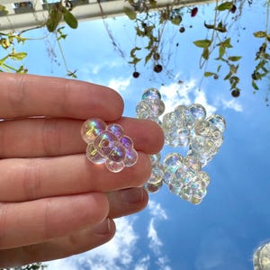 May include: A cluster of clear, iridescent glass beads, held in a hand, reflecting the sky. The beads are clustered together, resembling bubbles. The background shows a blue sky with clouds and greenery.