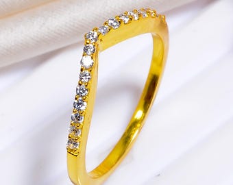 May include: A gold-colored V-shaped ring with a row of small, clear gemstones set along the top edge. The ring is displayed on a white surface with soft, blurred floral accents in the background. The ring's design is simple and elegant.