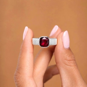 May include: A silver ring featuring a cushion-cut deep red gemstone set in a bezel. The band of the ring has a distinct vertical ribbed or grooved texture that extends around the sides, providing a structured and metallic appearance to the piece.
