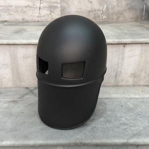 Può includere: Un casco nero opaco con una parte superiore arrotondata e una visiera. Presenta due aperture rettangolari per gli occhi. Il design del casco è minimalista e moderno, adatto a vari usi. Misure in centimetri.