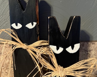 May include: Two black wooden cat figures with white painted eyes and raffia bows. The cat figures are in a rustic style, perfect for Halloween or fall decor. The figures are of different sizes.