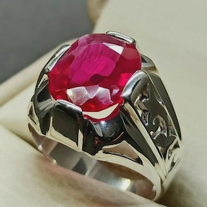 Könnte beinhalten: Ein silberner Ring mit einem großen, facettierten, oval geschliffenen Rubin. Der Ring hat ein detailliertes, durchbrochenes Design an den Seiten, und der Rubin hat einen leuchtenden Rotton. Der Ring ist vor einem neutralen Hintergrund platziert.