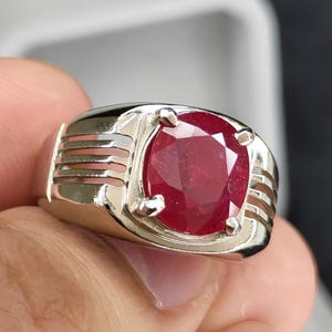 May include: A silver ring featuring a large, oval-cut ruby gemstone. The ruby is a deep red color and is set in a polished silver band with a classic design. The ring is a statement piece.