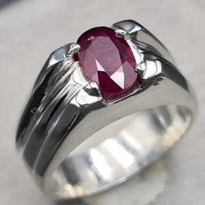 May include: A silver ring featuring a large, oval-cut ruby gemstone. The ruby is a deep, rich red color and is set in a polished silver band. The ring has a classic, elegant design, suitable for various occasions.