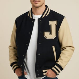 May include: A black and beige varsity jacket featuring a large letter "J" on the chest. The jacket has beige sleeves, a black body, and striped cuffs and collar. The jacket is buttoned up and worn over a white t-shirt.