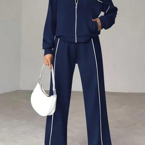 Può includere: Completo tuta blu navy con finiture bianche. Il set include una giacca con zip e pantaloni a gamba larga. Una borsa bianca è tenuta in mano. L'outfit è abbinato a scarpe da ginnastica bianche.