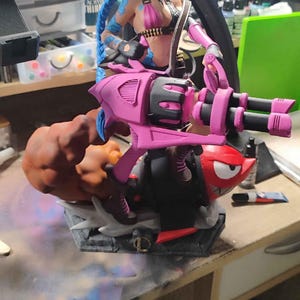 May include: A detailed figurine of a female character with blue hair, wearing a pink and black outfit, and holding a large pink weapon. The figure is mounted on a base with a cartoonish design, featuring a red character and orange smoke effects.