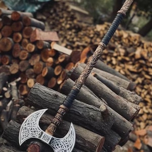 May include: A double-bladed axe with a silver blade and intricate Celtic knotwork design. The handle is wrapped in black leather and features carved wooden accents. The axe rests on a pile of wooden logs, showcasing its craftsmanship.
