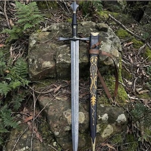 Könnte beinhalten: Ein zweischneidiges Stahlschwert mit schwarzem Griff und kreuzförmigem Parier, platziert neben einer schwarzen Scheide mit filigranen goldfarbenen Mustern und einem braunen Lederriemen zum sicheren Tragen der Waffe.
