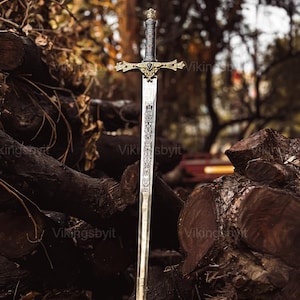 Op de afbeelding: Een middeleeuws zwaard met een sierlijke gouden handgreep en een zilveren lemmet, geplaatst op donkerbruine boomstammen. De afbeelding bevat de tekst "Vikingsbyit". Het zwaardontwerp suggereert historisch vakmanschap.