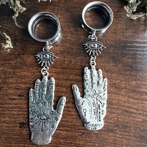 May include: A pair of silver-toned ear plugs featuring dangling charms. Each plug is attached to a small eye charm, which connects to a palmistry hand charm. The hands are engraved with various symbols, lines, and text including the words Life and Bracelet.